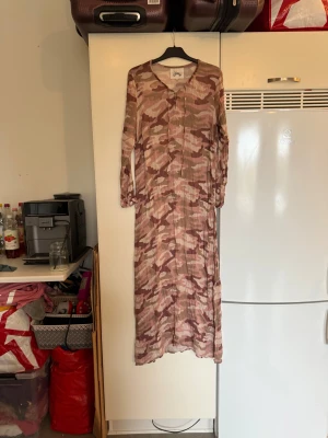Camouflage långklänning från Fira - Säljer en långklänning från Fira med coolt camouflage-mönster i beige, brun och rosa toner. Klänningen har v-ringad hals, knytband i midjan och långa ärmar med knappdetalj vid mudden. Materialet är lätt och luftigt, perfekt för dig som gillar avslappnad stil. Storlek L. Den ör i fint skick, men mer använd. Kan ha små tecken på användning. 