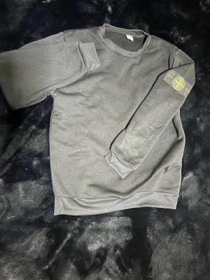 Stone Island grå och svart sweatshirt - Sweatshirt från Stone Island i både grått och svart, med klassisk rund hals och ribbade muddar. Ikonisk Stone Island patch på vänster ärm. Perfekt för dig som gillar stilrena och enkla plagg med streetkänsla.