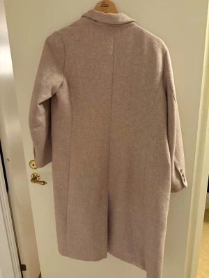 Beige ullkappa från H&M, stl 42 passar 40 st  - Snygg beige kappa från H&M i ullblandning, rak modell med klassisk krage och knappar vid ärmslut. Fodrad insida och flera fickor framtill. Perfekt för dig som vill ha en stilren och tidlös look.