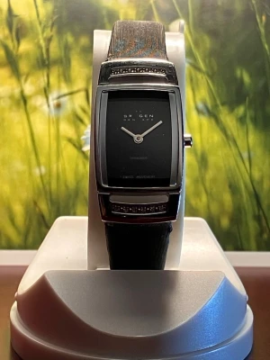 Svart Skagen klocka med diamantdetaljer - Snygg svart Skagen klocka med rektangulär urtavla och diskreta diamantdetaljer på boetten. Armbandet är svart och har en klassisk spänne i silverfärg. Urtavlan är minimalistisk med silverfärgade visare och en stilren look som passar till många outfits. Nytt, batteri, säljes i befintligt skick enligt bilderna!