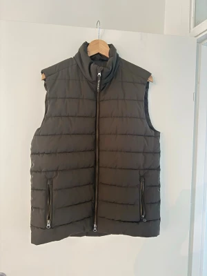 Mörkgrön dunväst från Bondelid - Snygg mörkgrön dunväst från Bondelid med quiltad design och hög krage. Västen har två fickor med dragkedja och en kraftig dragkedja framtill. Perfekt att slänga över en hoodie eller långärmad tröja för extra värme. Modern och stilren look.