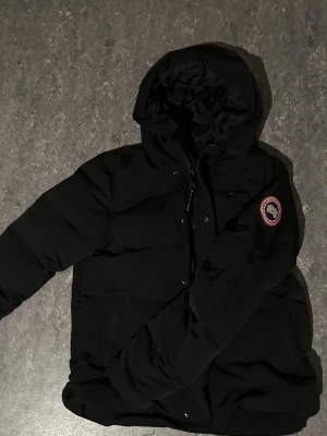 Svart pufferjacka från Canada Goose - Svart pufferjacka från Canada Goose med huva och klassisk logotyp på ärmen. Jackan har dragkedja med logotyp, tryckknappar och flera fickor. Perfekt för kalla dagar och har en clean, stilren look som funkar till streetwear. Materialet är syntet och dun.