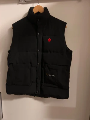Canada Goose Väst Dam - Svart dunväst från Canada Goose med hög krage, två stora fickor framtill och röd lönnbladsbroderi på bröstet. Västen har tryckknappar och dragkedja. Priset kan diskuteras vid snabb affär. Använd väldigt sparsamt men små defekter vid lapparna