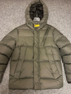 Grön PJS pufferjacka med huva - Snygg mörkgrön pufferjacka från Parajumpers med stor huva, gul detalj vid halsen och patch på ärmen. Jackan har flera fickor med dragkedja och quiltad design. Perfekt för kalla dagar och har en cool streetstil.