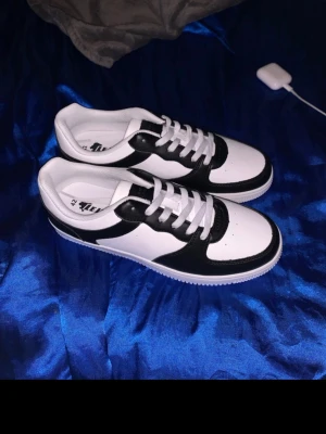 Svarta och vita sneakers från Lejon - Snygga svartvita sneakers från Lejon i storlek 42. Skorna har klassisk design med vit bas och svarta detaljer, rund tå och vita snören. Ovandelen är i syntetmaterial och sulan är vit med mönstrad undersida för bra grepp. Perfekta för en clean och stilren look.