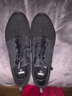 Svarta sneakers från Bench strl 42 - Säljer ett par helsvarta sneakers från Bench i storlek 42. Skorna har en stickad ovandel med snygga detaljer och paneler i syntet, samt snörning och en cool mönstrad sula. Perfekta för dig som gillar stilrena och sportiga skor.