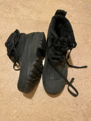 Svarta kängor med grov sula - Svarta kängor med högt skaft och grov, mönstrad sula. Skorna har snörning framtill och är tillverkade i syntetmaterial med en robust design som passar perfekt för kyliga dagar. Insidan är fodrad för extra komfort och värme.