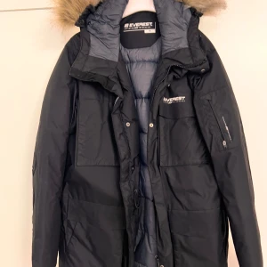 Svart parkasjacka Everest med fuskpäls - Svart parkasjacka från Everest med huva och fuskpäls. Jackan har flera stora fickor, dragkedja och knappar framtill samt logga på bröstet. Vadderad insida för extra värme, perfekt för kalla dagar. Klassisk och stilren design.