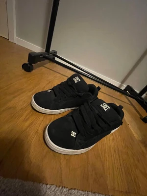 Svarta DC sneakers med vit logga - Säljer ett par klassiska DC sneakers i svart mocka med vit DC-logga på sidan och tungan. Skorna har tjocka svarta snören, platt vit sula och rund tå. Perfekta för dig som gillar skate-stil och vill ha ett par snygga och bekväma sneakers.