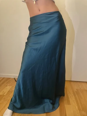 Mörkgrön satinkjol i maxilängd - Säljer en lång, mörkblå satinkjol med snygg glansig finish. Kjolen har en figurnära passform upptill och faller snyggt ner mot golvet. Perfekt för dig som gillar en elegant och stilren look. Kjolen är helt utan mönster och har en mjuk, följsam känsla.