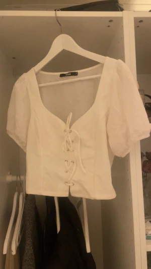 Vit topp med snörning från Bik Bok - Supersöt vit blus från Bik Bok med puffiga korta ärmar och snygg snörning framtill. Croppad passform och v-ringad hals ger en trendig look. Materialet känns lätt och luftigt, perfekt för varmare dagar. Enkel att matcha med både jeans och kjol.