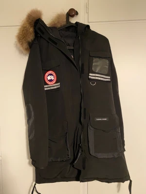 Svart parkajacka från Canada Goose - Svart parkajacka från Canada Goose med päls på huvan, flera fickor och reflekterande detaljer. Jackan har klassisk logga på ärmen, dragkedja framtill och är designad för att hålla dig varm under kalla dagar. Perfekt för dig som vill ha en stilren och funktionell vinterjacka.