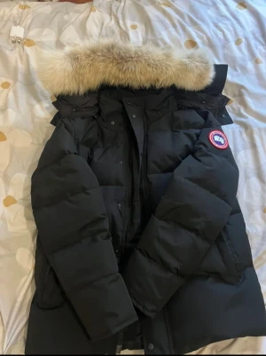 Svart Canada Goose dunjacka med päls - Säljer en svart dunjacka från Canada Goose med ikonisk röd och vit patch på ärmen. Jackan har en stor fluffig päls på huvan, dragkedja och knappar framtill samt långa ärmar. Perfekt för kalla vinterdagar och riktigt snygg streetstil.