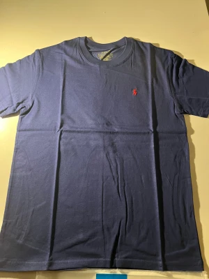 Marinblå t-shirt Polo Ralph Lauren - Snygg marinblå t-shirt från Polo Ralph Lauren med klassisk röd broderad logga på bröstet. Modellen är custom slim fit med rund hals och korta ärmar. Tillverkad i mjuk 100% bomull, perfekt för en clean och stilren look.