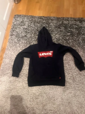 Marin blå Levi's hoodie med röd logga - Marin blå hoodie från Levi's med stor röd och vit logga på bröstet. Klassisk modell med huva och känguruficka framtill. Mjuk insida och ribbade muddar vid ärmslut och nederkant. Perfekt för dig som gillar streetwear och enkel stil.