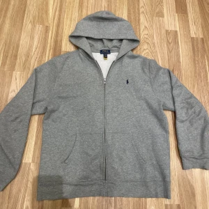 Grå Zip hoodie Ralph Lauren XS/S - Grå hoodie från Ralph Lauren i storlek XL barn men passar XS och Small i herr. Dragkedja, luva och två fickor. Blå logga på bröstet.