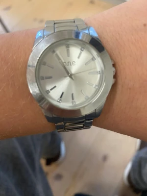 Silverfärgad klocka från One - Stilren klocka från One med rund urtavla i silver och matchande länkarmband i metall. Urtavlan har minimalistiska markeringar och en diskret logga. Perfekt för dig som gillar enkel och modern design.