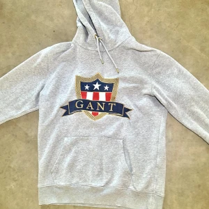 Grå hoodie med broderad GANT-logga - Snygg grå hoodie från GANT med stort broderat emblem i blått, rött, vitt och guld på bröstet. Tröjan har huva med snörning och guldfärgade detaljer, samt en klassisk känguruficka framtill. Perfekt för dig som gillar sportig och stilren streetwear. Köpt i GANT butiken i Göteborg men har inget kvitto kvar. 🫣Köpt för 800kr🫣 🍀Säljer för 400🍀Den är strl M men du som har L och vill ha en liten slimmare tröja funkar också perfekt!