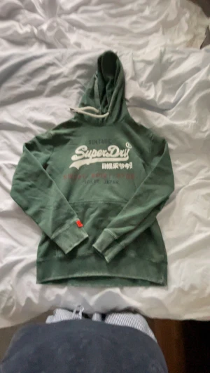 Grön Superdry hoodie med tryck - Mörkgrön hoodie från Superdry med vit logga och japanska tecken på bröstet. Tröjan har huva med snörning, känguruficka och långa ärmar. Mjuk insida och klassisk passform, perfekt för chill dagar.