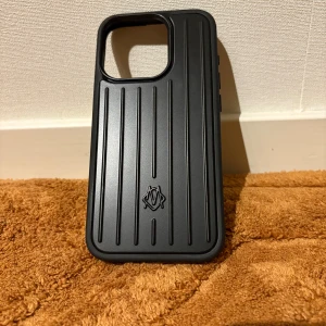 Rimowa iPhone 15 Pro Case - Elegant och robust Rimowa-skal för iPhone 15 Pro, tillverkat i Tyskland. Svart design med ikoniska Rimowa-ränder och logotyp. Skalet är i mycket gott skick utan synligt slitage, levereras med originalförpackning.