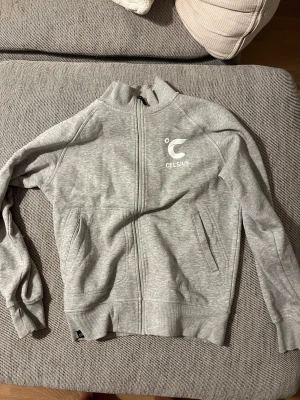 Grå ziptröja från Celsius - Snygg grå ziptröja från Celsius med logga på både bröstet och ryggen. Tröjan har ribbade muddar, ståkrage och två fickor framtill. Perfekt för träning eller chill, med mjukt material och stilren look. Svår att få tag på.