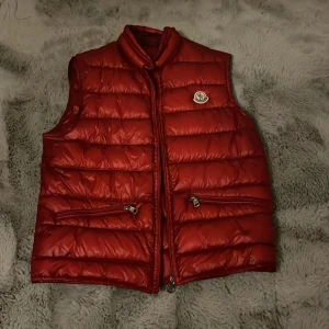 Röd dunväst från Moncler (ÄKTA) - Säljer en röd dunväst från Moncler med klassisk quiltad design och två dragkedjefickor framtill. Västen har hög krage och Moncler-logga på bröstet. Perfekt att slänga över en hoodie eller tröja för extra värme med stil. Västen har inga defekter eller hål, men de syns att den har varit använd. Har tyvärr inte kvar original påsen men de fins en kod på laparna där man kan skriva in och checka. Priset är diskuterbart, hör av er vid frågor // Leo
