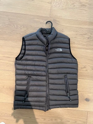 Grå dunväst från The North Face - Snygg grå dunväst från The North Face med quiltad design och hög krage. Västen har två praktiska fickor med dragkedja på utsidan och en innerficka med dragkedja. Perfekt att slänga över en hoodie för extra värme. Logga på bröstet och insidan.