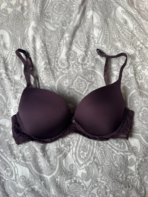 Mörklila push-up bh från Lindex  - Snygg mörklila push-up bh från Lindex med släta kupor och spetsdetaljer längs sidorna. Justerbara axelband och klassisk hakknäppning bak. Materialet är mjukt och skönt mot huden, perfekt för dig som gillar en feminin look med lite extra stöd.