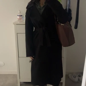 Svart lång kappa med bälte - Säljer en svart lång kappa med bälte i midjan och klassisk krage. Kappan har långa ärmar och en stilren, tidlös look som passar perfekt till höst och vinter. Enkel att matcha med olika outfits och ger en snygg siluett.