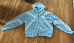 Ljusblå hoodie med vita detaljer rare humans - Säljer en ljusblå hoodie från rare humans med vita grafiska detaljer på ärmar och bröst. Tröjan har huva, dragkedja framtill och två fickor. Materialet är mjuk bomull och passformen är loose, perfekt för chill dagar. Långärmad och riktigt snygg design!