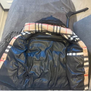 Svart dunväst från Burberry, L - Svart dunväst från Burberry i storlek L med klassiskt rutigt foder i beige, rött och svart. Västen har dragkedja och tryckknappar framtill samt två fickor med dragkedja. Materialet är polyester och dun, perfekt för kyliga dagar. Den ikoniska Burberry-looken syns tydligt i fodret.