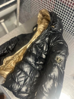 Svart och guld pufferjacka från Rockandblue - Säljer en riktigt snygg svart pufferjacka från Rockandblue med glansig finish och guldfärgat innerfoder. Jackan har huva med pälsdetaljer, två dragkedjefickor och Rockandblue-logga på ärmen. Perfekt för dig som vill sticka ut i vinterkylan.