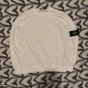 Vit sweatshirt från Stone Island - Vit långärmad sweatshirt från Stone Island med klassisk rund hals och ikonisk logotyp-patch på vänster ärm. Tröjan är i mjuk bomull och har en clean, stilren look som passar perfekt till jeans eller cargo. Taggarna sitter kvar och passformen är normal.