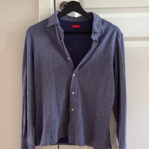 Blå långärmad skjorta från Altea - Snygg blå skjorta från Altea med klassisk krage och knappar framtill. Skjortan har lång ärm och coola mörkblå armbågslappar som ger extra stilpoäng. Materialet är mjukt och känns som bomull, perfekt för en chill och avslappnad look.