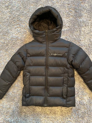 Svart dunjacka från Sail Racing - Svart dunjacka från Sail Racing med huva och stilren logga på bröstet. Jackan har quiltad design, dragkedja framtill och är tillverkad i polyester med dunfyllning. Perfekt för kalla dagar och har en clean look som funkar till det mesta.