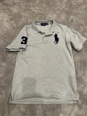Ralph Lauren Polo Shirt - Snygg grå pikétröja från Polo Ralph Lauren i custom slim fit. Tröjan har korta ärmar, klassisk krage, stor marinblå logga på bröstet och en broderad siffra 3 på ärmen. 