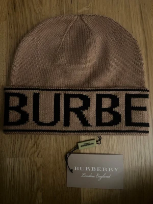 Burberry Mössa - Snygg beige mössa från Burberry med stor svart logotyp runt uppviket. Mössan är stickad och har en klassisk, rundad form. Perfekt för dig som vill ha en stilren accessoar med lyxig känsla och tydlig branding.