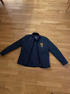 Morris quiltad jacka marinblå S - Quiltad marinblå jacka från Morris med snyggt broderat emblem på bröstet. Jackan har lång ärm, ståkrage och dragkedja framtill. Klassisk rutig quiltning och sportig look, perfekt för dig som gillar stilren design.