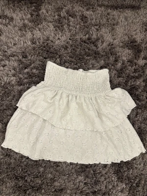 Vit volangkjol från Gina Tricot - Supersöt vit kortkjol från Gina Tricot med broderade blommor och bred smock i midjan. Kjolen har dubbla volanger och är gjord i ett lätt bomullstyg som känns perfekt för varma dagar. Enkel att matcha med crop tops eller en oversized hoodie.