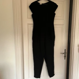 Svart byxdress från Monsoon - Stilren svart jumpsuit från Monsoon i storlek S/36-38. Modellen är tidlös med korta ärmar, fickor framtill. Byxan avsmalnande och slutar i ankelhöjd. Snyggt skäddad midja. Materialet känns mjukt och följsamt. Dragkedja i ryggen. Funkar snyggt till både klack, sandal, sneakers och stövlar. Styla med skärp eller mer loose fit. 