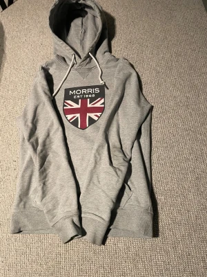 Grå hoodie Morris XS  - Snygg grå hoodie från Morris i storlek XS. Snyggt tryck med texten 'Morris Est 1968' på bröstet. Tröjan har huva med snörning, ribbade muddar och känguruficka. Perfekt för dig som gillar klassisk stil med en twist. Hör 