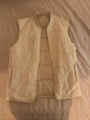 Väst - Snygg beige dunväst från Zara med quiltad design och ståkrage. Västen har en diskret bröstficka med dragkedja och är perfekt att slänga över en hoodie eller tröja. Lätt och stilren look som funkar till många outfits.