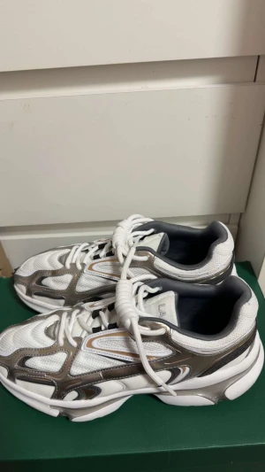 Lacoste vita sneakers med silverdetaljer - Snygga vita sneakers från Lacoste med silvriga och grå detaljer. Ovandelen är i mesh och syntet, med coola mönstrade paneler och rund tå. Skorna har platt sula och klassisk snörning. Perfekt för dig som gillar sportig och modern stil.