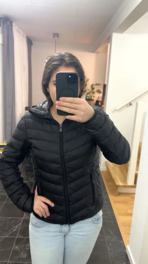 Svart pufferjacka från McKinley - Svart pufferjacka från McKinley med huva och dragkedja framtill. Jackan har två fickor med dragkedja och quiltad design. Perfekt för kyliga dagar och enkel att matcha med allt. Snygg och stilren modell som håller värmen.