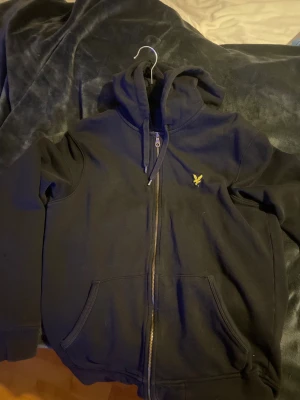 Svart hoodie från Lyle & Scott - Svart hoodie från Lyle & Scott i storlek M. Tröjan har dragkedja framtill, huva med snörning och två fickor. Klassisk gul Lyle & Scott-logga på bröstet. Perfekt för chill dagar och enkel att matcha med jeans eller joggers.