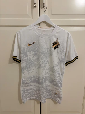 Vit AIK fotbollströja Nike - AIK fotbollströja från Nike i vit med grått grafiskt mönster över hela framsidan. Guld- och svartdetaljer på ärmsluten, AIK-emblem på bröstet och Nike-logga i guld. Materialet är lätt och andas, perfekt för match eller träning.