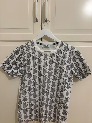 Vit mönstrad t-shirt från Lacoste - Säljer en vit t-shirt från Lacoste med svart grafiskt mönster över hela plagget. Klassisk rund hals och korta ärmar. Materialet är mjuk bomull och den har den ikoniska gröna krokodilen broderad på bröstet. Perfekt för dig som gillar stilrena detaljer.