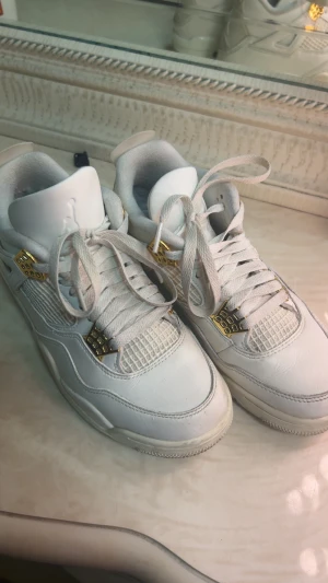 Nike Air Jordan 4 vit med gulddetaljer - Nike Air Jordan 4 sneakers i helvit skinn med snygga gulddetaljer vid snörningen. Klassisk siluett med rund tå, platt sula och meshpaneler på ovansidan. Jumpman-logga på hälen och tungan. Perfekt för dig som vill ha en clean och exklusiv look.