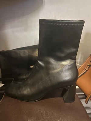 .Svarta stövletter med klack och dragkedja - Säljer ett par aldrig använda eleganta svarta stövletter i skinn med rund tå och stabil klack. Stövletterna har dragkedja på insidan för smidig på- och avtagning. Den stilrena designen med slät yta och diskreta sömmar gör dem perfekta för en minimalistisk och tidlös stil. Skorna köptes för ett år sedan men har endast legat i garderoben sedan dess och är i nyskick.