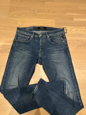 Replay Anbass blå jeans slim fit - Snygga Replay Anbass jeans i klassisk blå tvätt med lätt slitning och smal passform.  L32. Ett litet hål finns på sidan om jeansen, och en en hälla är trasig men inget märkvärdigt. Modellen har fem fickor, dragkedja och knapp, samt Replay-logga på myntfickan. Perfekt för dig som gillar en modern och stilren look. 